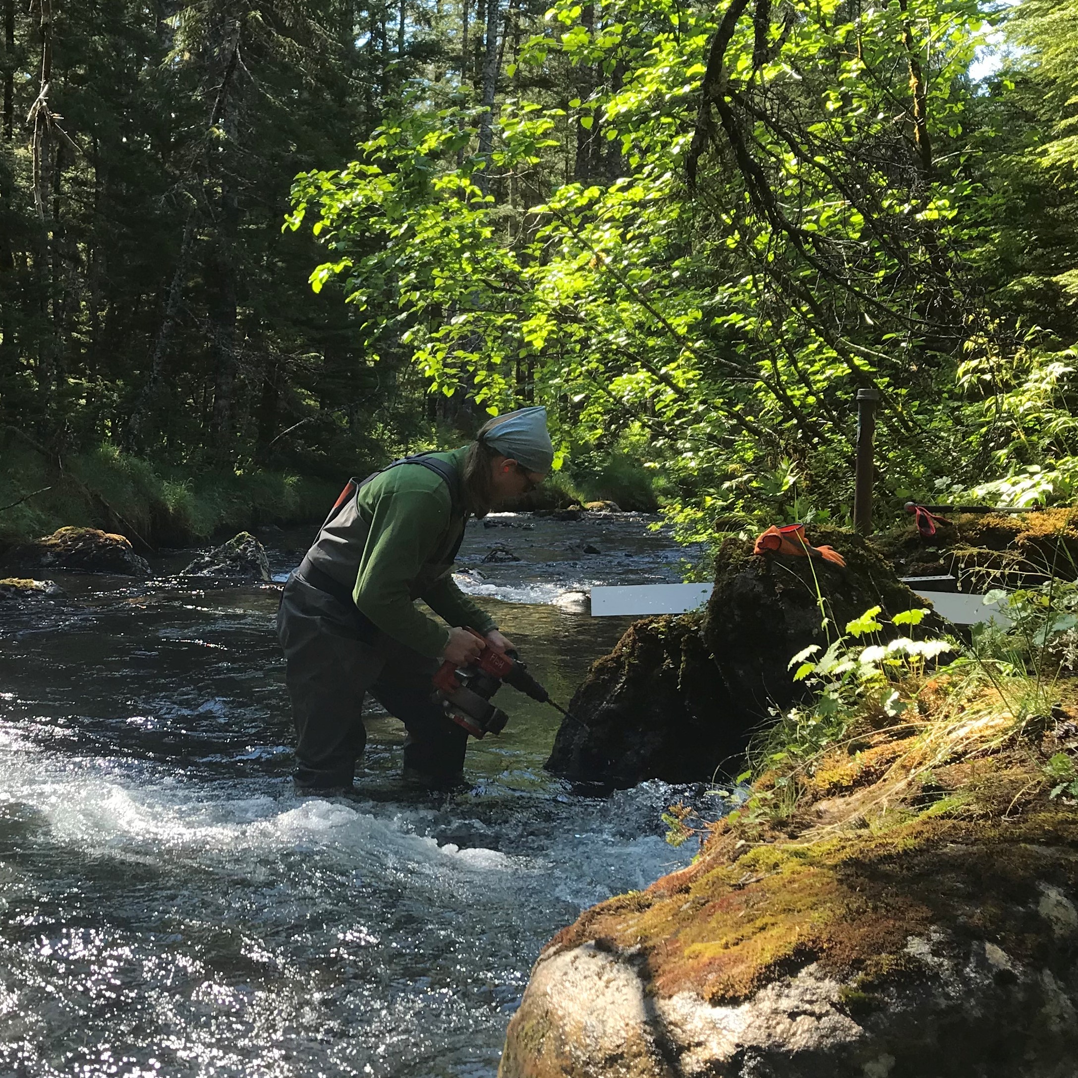 WRS student Dean Anderson is standing in a fast paced river with a drill next to a stone. He is drilling a measuring device into the rock.